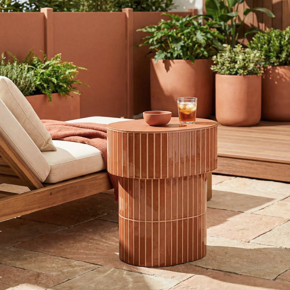 An oval outdoor end table with a glossy brown ceramic finish with grooved detailing, styled next to a lounge chair on a patio with potted plants in the background.