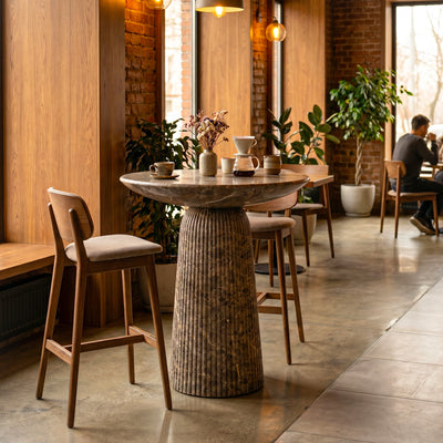 A round concrete counter-height bar table in a faux emperador marble finish with a fluted pedestal base in a bright, modern cafe setting. 