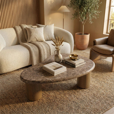 A faux emperador marble top with faux wooden legs coffee table in a styled living room. Styled with a small vase, books, and a gold bowl, set on a woven jute rug next to a cream boucle sofa. 